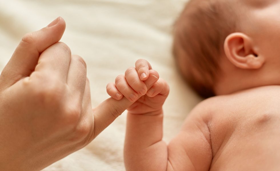 Faceless mother with naked baby, infant holding mommy's finger, mum spending time with her tiny child on light background.